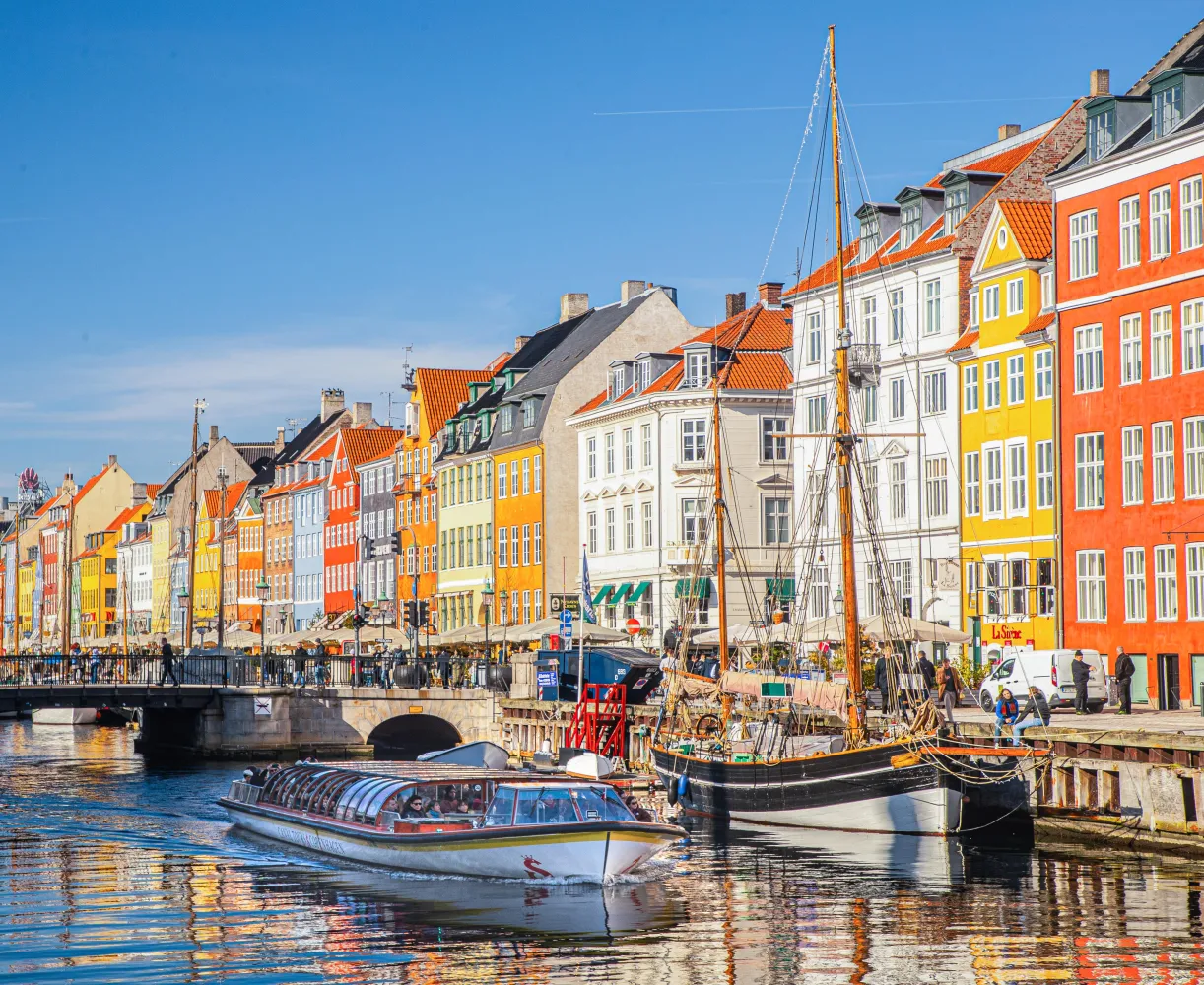 Colorful historic buildings line the Nyhavn waterfront in Copenhagen on a sunny day, with a sightseeing boat gliding through the canal and a docked wooden sailboat in the foreground.